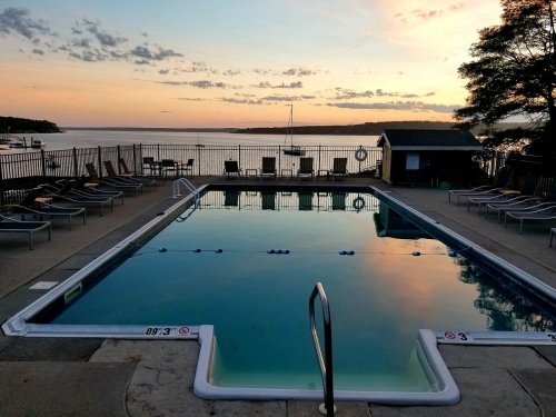A serene pool at sunset, reflecting the sky, with lounge chairs and a lake in the background.
