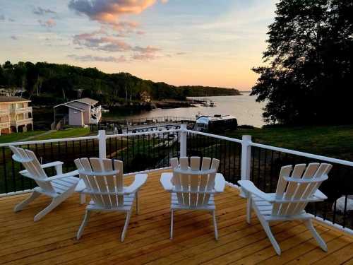 A serene lakeside view at sunset, featuring white Adirondack chairs on a wooden deck overlooking the water and boats.