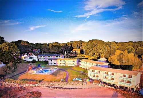 Aerial view of a beachside motel with a pool, surrounded by trees and grassy areas under a colorful sky.