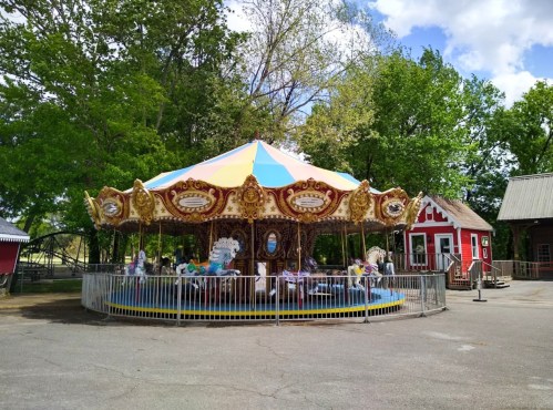 A colorful carousel with ornate decorations, surrounded by trees and a small red building in the background.