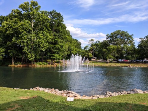 A serene pond with a fountain surrounded by lush green trees and a clear blue sky.