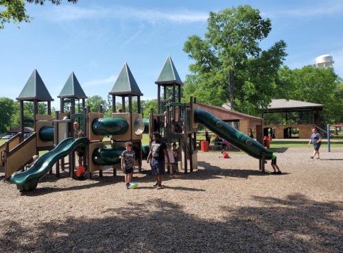 Children playing on a colorful playground with slides and climbing structures, surrounded by trees and a clear blue sky.