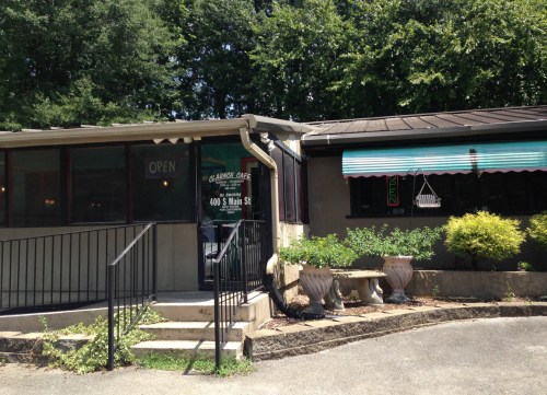 A small restaurant with an open sign, featuring a ramp, outdoor seating, and green awnings on a sunny day.