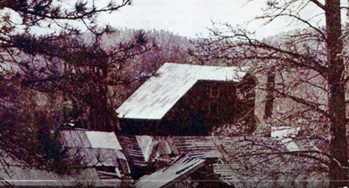 A damaged building surrounded by trees, with a partially collapsed roof and a mountainous background.