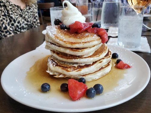 A stack of fluffy pancakes topped with whipped cream, blueberries, and strawberries, drizzled with syrup.