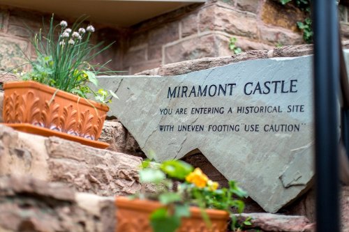 Sign at Miramont Castle warns visitors of uneven footing, surrounded by potted plants on stone steps.