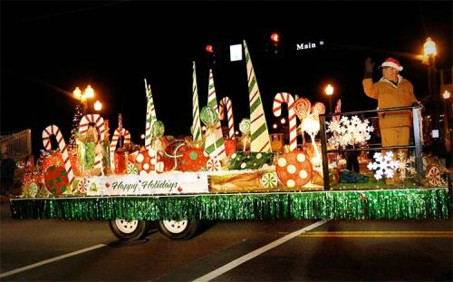 A festive parade float decorated with candy canes, ornaments, and a "Happy Holidays" sign, illuminated at night.
