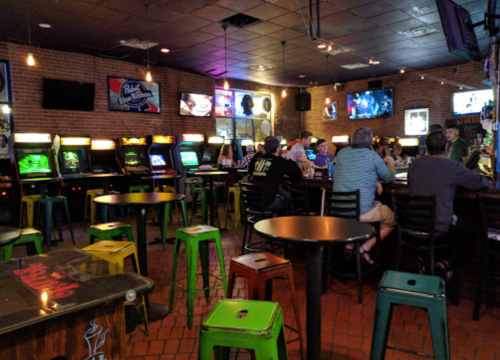 A lively bar with arcade games, colorful stools, and patrons enjoying drinks and entertainment on screens.