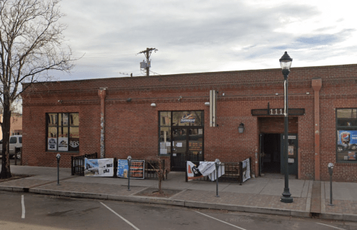 A brick building with large windows, featuring outdoor seating and banners, set against a cloudy sky.