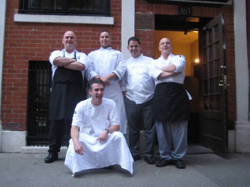 Five chefs pose together outside a restaurant, smiling and wearing white and black uniforms.