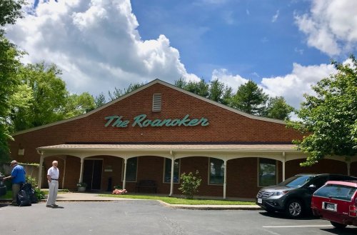 A brick building with "The Roanoker" sign, surrounded by trees and a blue sky with clouds. Two people stand outside.