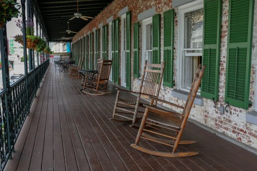 A porch with wooden rocking chairs and green shutters, featuring a rustic brick wall and a wooden floor.
