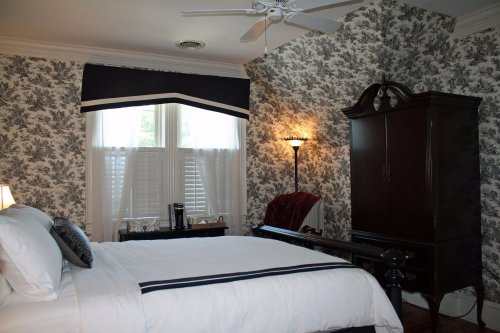 Cozy bedroom with floral wallpaper, a bed, a dresser, and a window with blinds, featuring soft lighting.