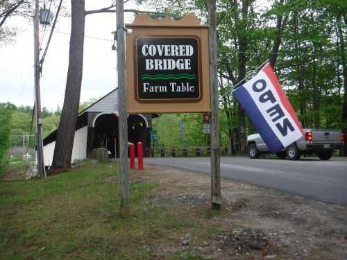 Sign for "Covered Bridge Farm Table" with a flag nearby, set in a rural area with a covered bridge in the background.