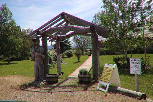A wooden archway with a carved figure, leading to a museum, surrounded by greenery and parking signs.