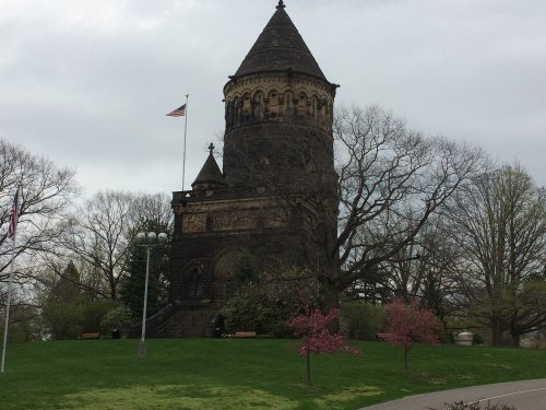 Historic stone tower with a conical roof, surrounded by trees and blooming flowers, under a cloudy sky.