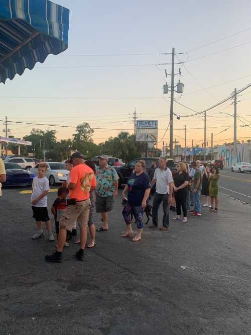 A line of people waiting outside a store at sunset, with cars parked along the street.