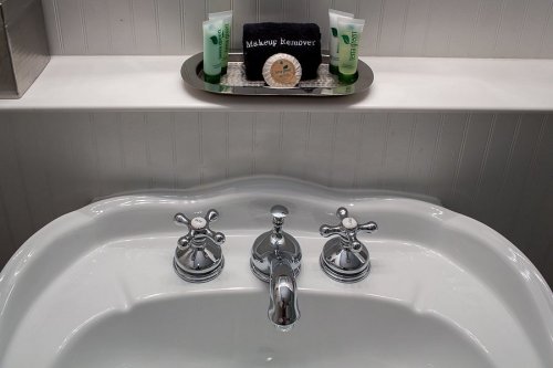A white sink with chrome faucets, featuring skincare products and a towel on a shelf above.