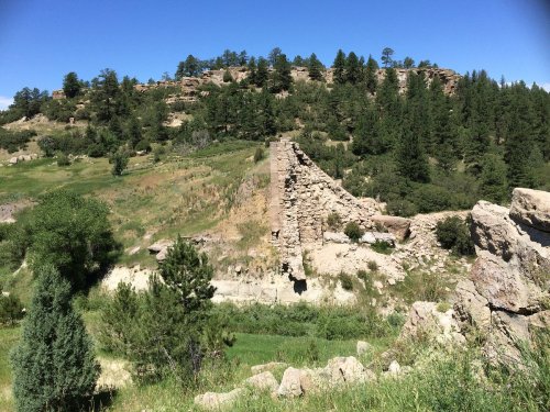 Ruins of a stone structure on a hillside, surrounded by greenery and trees under a clear blue sky.