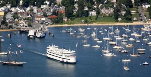 Aerial view of a harbor filled with sailboats, a ferry, and a coastal town with houses and trees in the background.