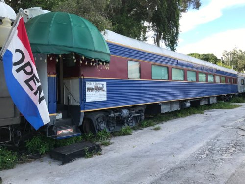 A vintage train car with a green awning, featuring an "Open" flag and a sign on the side, set in a grassy area.