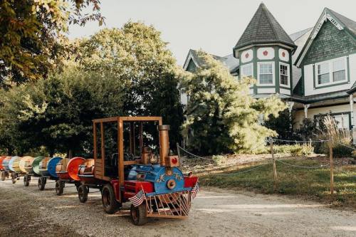 A colorful miniature train parked on a gravel path near a large, charming house surrounded by trees.