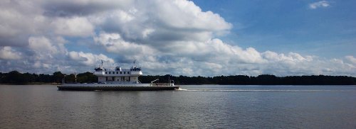 A ferry glides across a calm river under a blue sky with fluffy clouds. Lush greenery lines the shore.