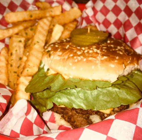 A sesame seed burger with lettuce and a pickle, served with crinkle-cut fries in a red and white checkered basket.