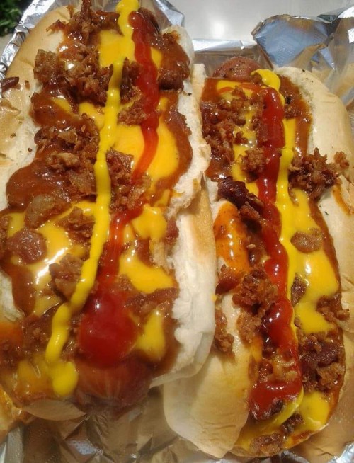 Two loaded hot dogs topped with mustard, ketchup, and crispy bacon bits, served on a foil tray.