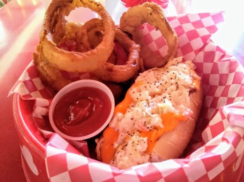 A basket with a hot dog topped with cheese and onions, served with onion rings and a side of ketchup.