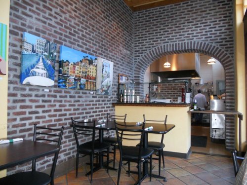 Cozy restaurant interior with brick walls, artwork, and a view of the kitchen. Black tables and chairs are set for dining.