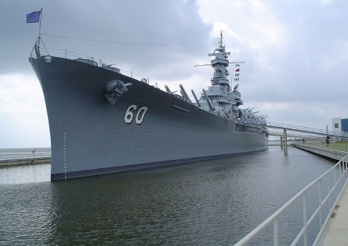 A large battleship docked in calm waters, with a walkway leading to it and flags flying on the mast.