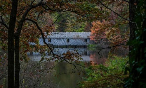 A rustic wooden bridge house surrounded by autumn foliage and reflecting in a calm pond.