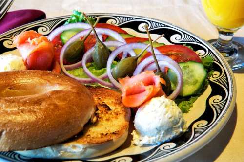 A plate with a toasted bagel, cream cheese, fresh salad with tomatoes, cucumbers, onions, and capers, alongside a glass of juice.