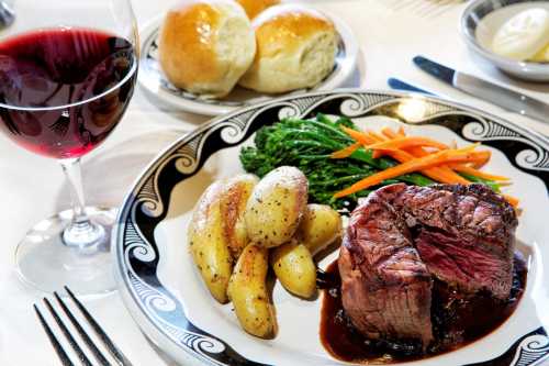 A plate of steak with roasted potatoes, carrots, and greens, accompanied by a glass of red wine and dinner rolls.