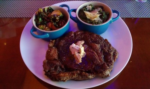 A large steak topped with butter, served with two bowls of mixed vegetables and greens on a white plate.