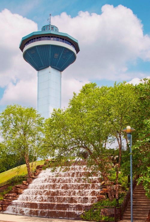 A tall observation tower stands above a cascading waterfall, surrounded by lush green trees and a blue sky.