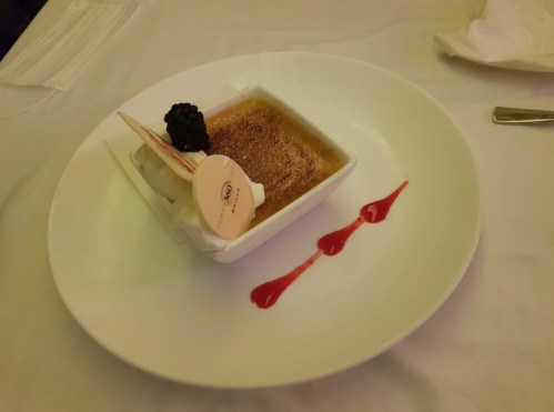 A creamy dessert in a square dish, topped with a black berry and drizzled with red sauce on a white plate.