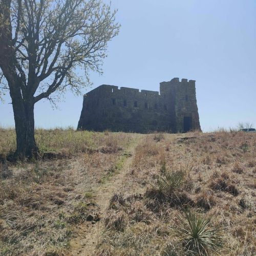 A weathered stone structure stands on a grassy hill, surrounded by sparse trees and open land under a clear blue sky.