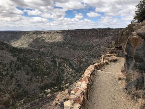 A rocky cliffside path overlooks a vast, green valley under a cloudy sky.
