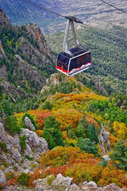 A cable car glides over vibrant autumn foliage and rocky terrain in a mountainous landscape.