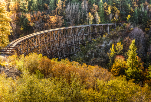 A wooden trestle bridge arches over a colorful autumn landscape with trees in shades of orange, yellow, and green.