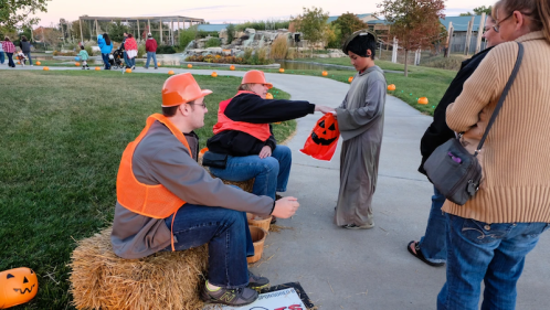 Two people in orange vests sit on hay bales while a child in a costume receives candy from them. Halloween decorations are visible.