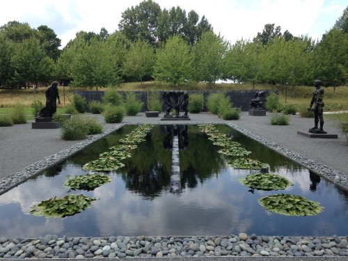A serene garden with a reflective pond, surrounded by sculptures and lush greenery under a cloudy sky.