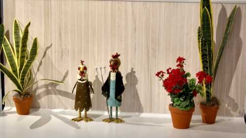 Two whimsical metal chicken figurines stand between potted plants and red geraniums against a light wooden background.