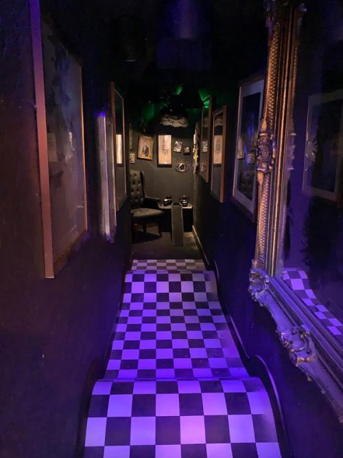 A dimly lit hallway with checkered flooring, framed artwork, and a chair at the end, creating a mysterious atmosphere.