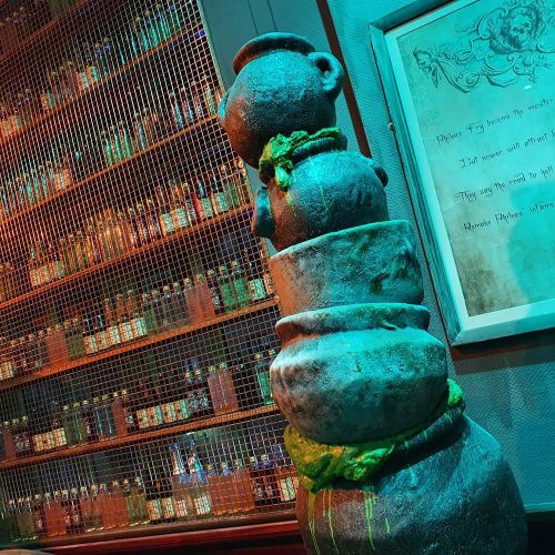 A stack of stone pots with green moss, set against a backdrop of shelves filled with colorful bottles.
