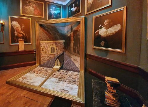 A 3D art installation in a gallery, featuring a framed scene of a stone building and a sloped roof, surrounded by portraits.