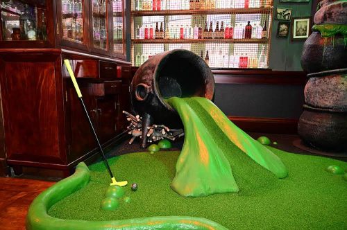 A whimsical mini-golf scene featuring a giant cauldron spilling green liquid onto a grassy surface with a golf club nearby.