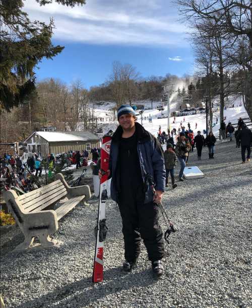 A skier stands on a gravel path holding skis, with a snowy slope and people in the background on a sunny day.